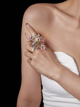 A person wearing a white strapless top holds CAMELLIA's Libra Flower Ring, featuring a handcrafted floral arrangement with a red heart charm, on their shoulder against a black background.